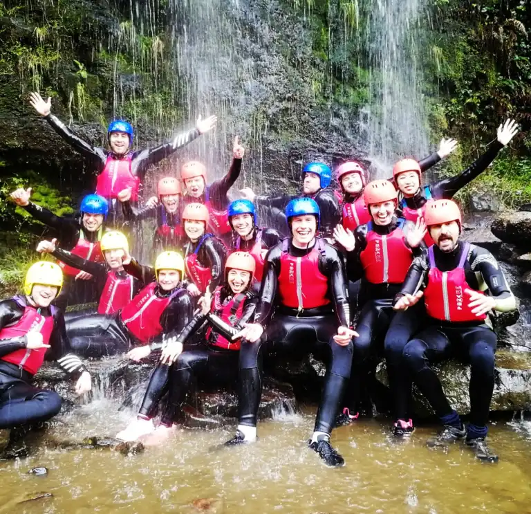 A group of people wearing wet suits, helmets and life jackets under a waterfall.