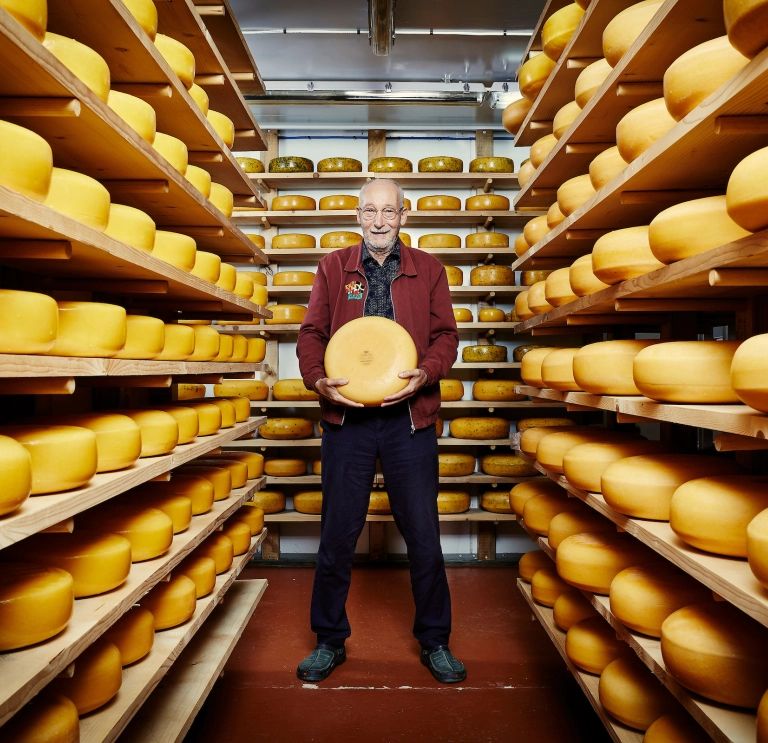 The owner holding a block of cheese surrounded by cheese on shelves.
