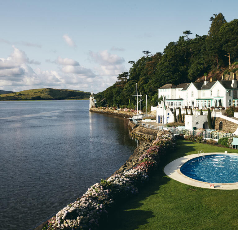 A white-fronted hotel and outdoor swimming pool by a river.