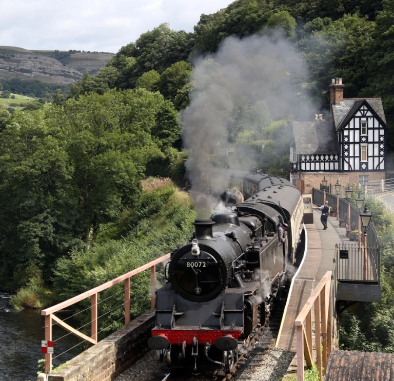 A steam train pulling out of a station alongside a lake.