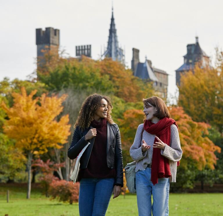 Two women walking in Autumn through Bute Park with trees and castle in the background