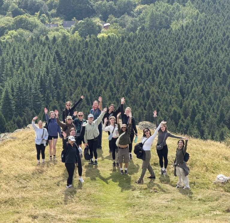 Group of people standing on a grassy hilltop with arms raised, overlooking a vast forest of evergreen trees and scattered countryside houses in the distance