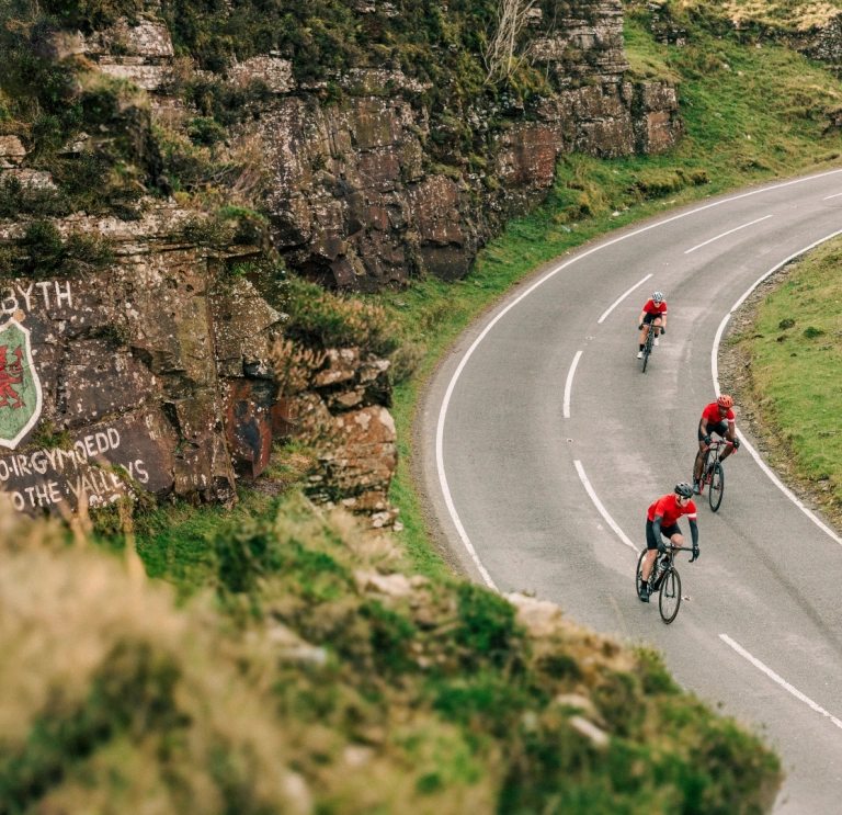 Cyclists riding along a winding mountain road in Wales with a painted rock face beside them.