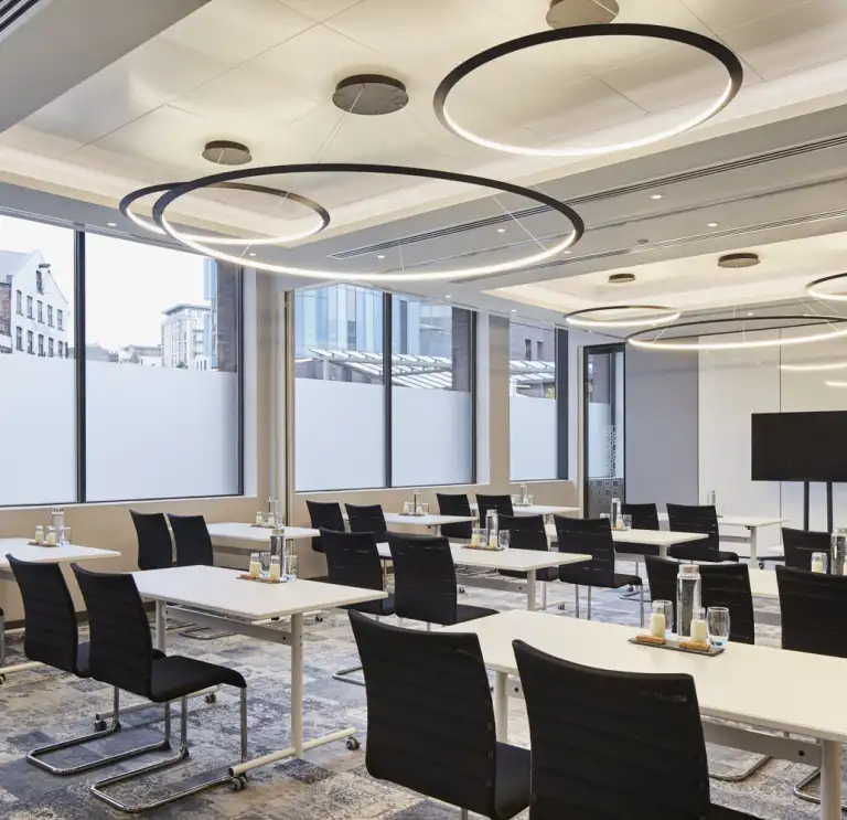 Meeting room featuring modern tables and chairs, large windows with city views, and circular ceiling lights, set up for a professional corporate event or training session.