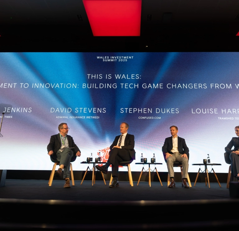 fPanel discussion on stage at a conference, with four speakers seated beneath a large screen reading “This is Wales: Investment to Innovation – Building Tech Game Changers from Wales.”