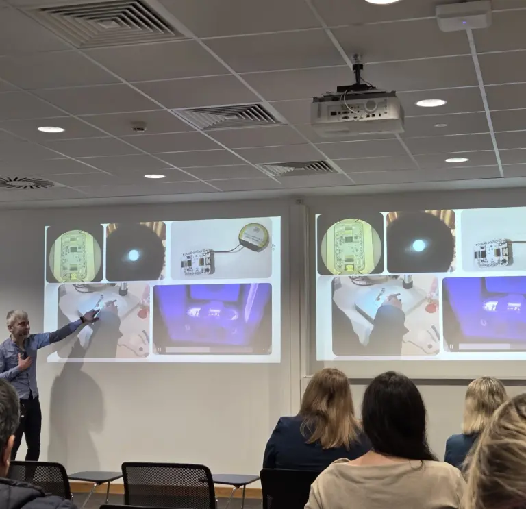 Presenter pointing to projected slides showing close‑up technical images, with an audience seated in a lecture room