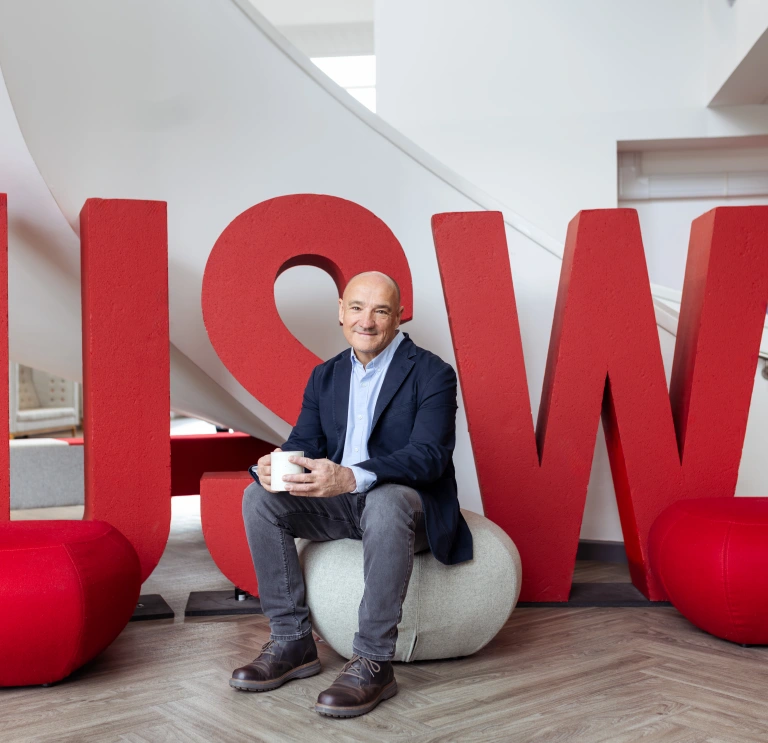Person seated on a stool holding a mug, in front of large red “USW” letters in a modern indoor space.