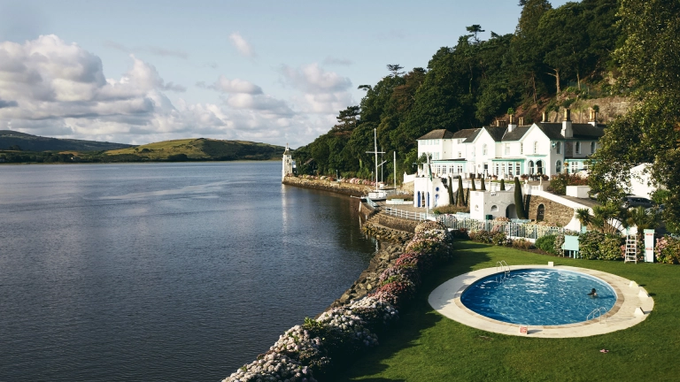 A white-fronted hotel and outdoor swimming pool by a river.