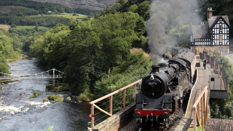 A steam train pulling out of a station alongside a lake.