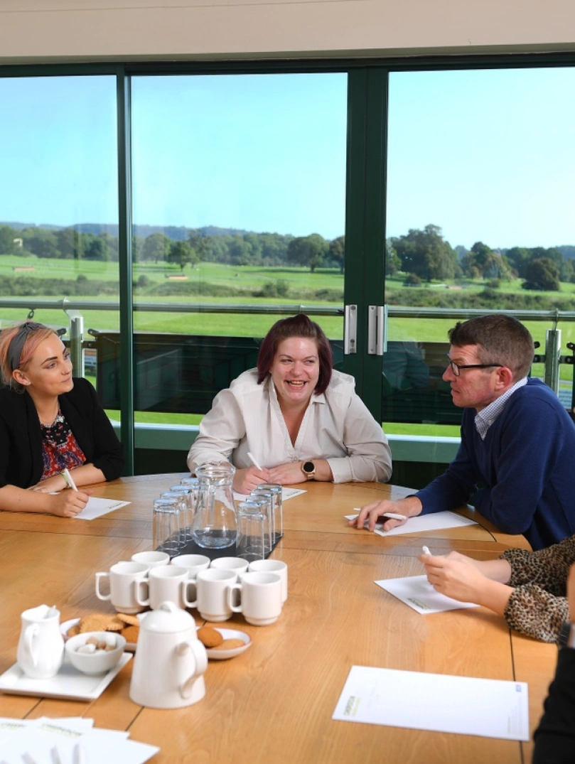 People around a meeting table with views of a racecourse beyond.