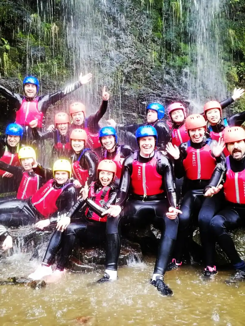 A group of people wearing wet suits, helmets and life jackets under a waterfall.