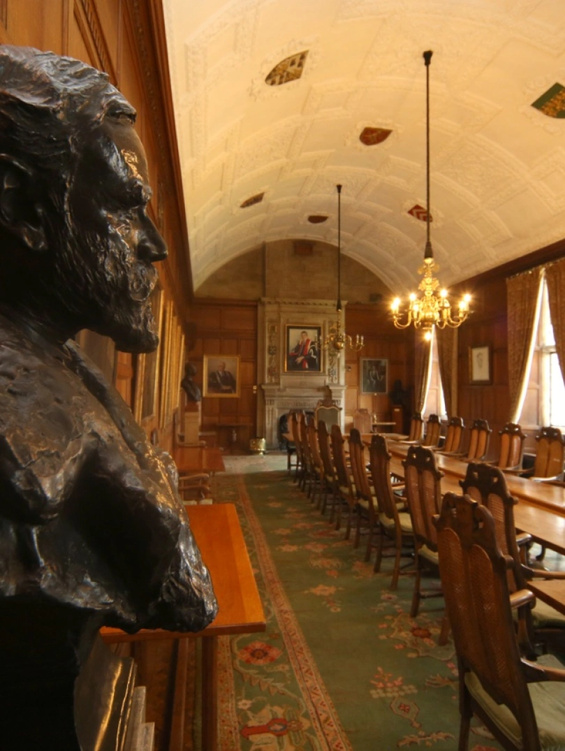 The council chamber witha head statue of a man low chandelier lights over a log table with fireplace at the end.