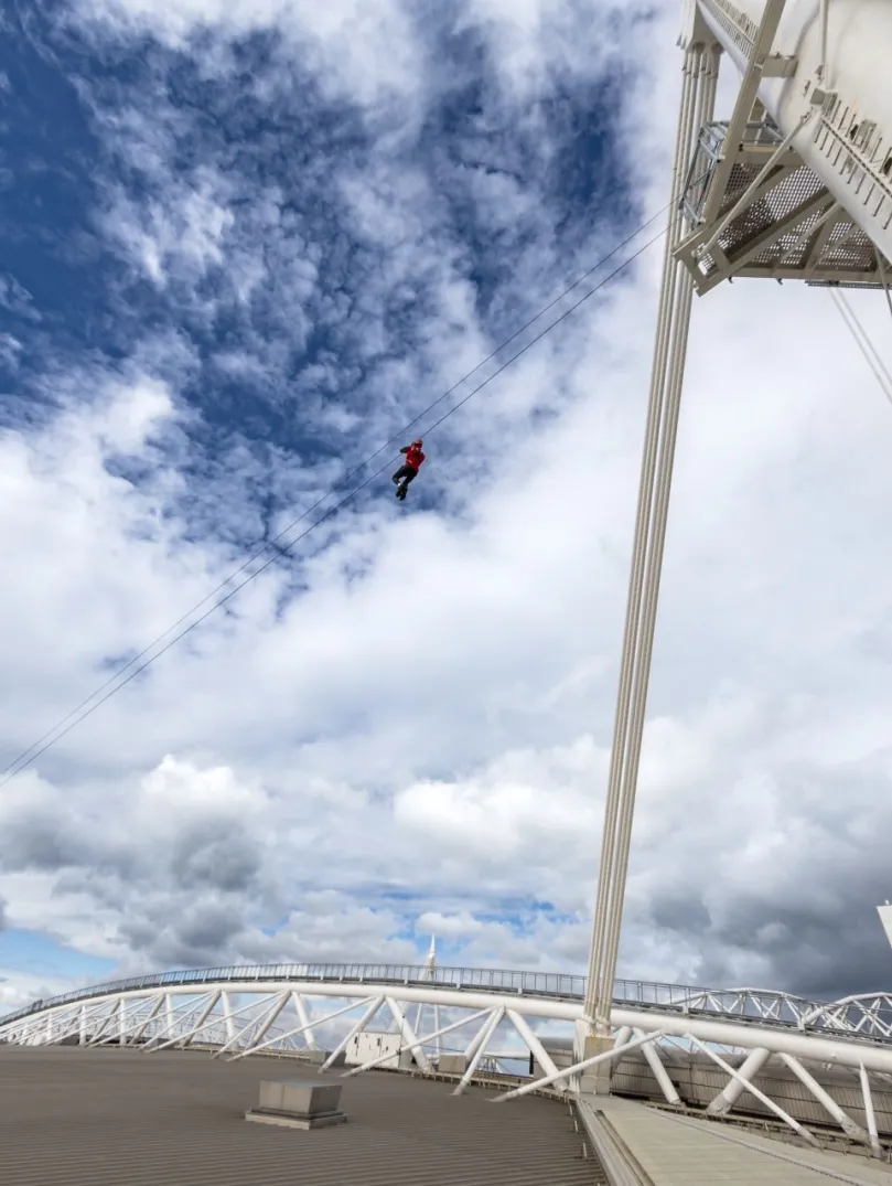 A person ziplining above the roof of a sports stadium.