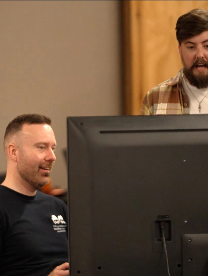 Ambassador Richard Hebblewhte sitting at a computer screen talking with a collegue standing behind him in a checked shirt