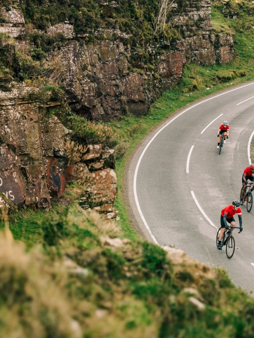Cyclists riding along a winding mountain road in Wales with a painted rock face beside them.