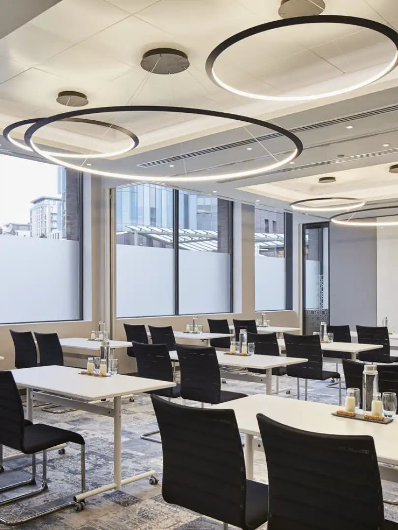 Meeting room featuring modern tables and chairs, large windows with city views, and circular ceiling lights, set up for a professional corporate event or training session.