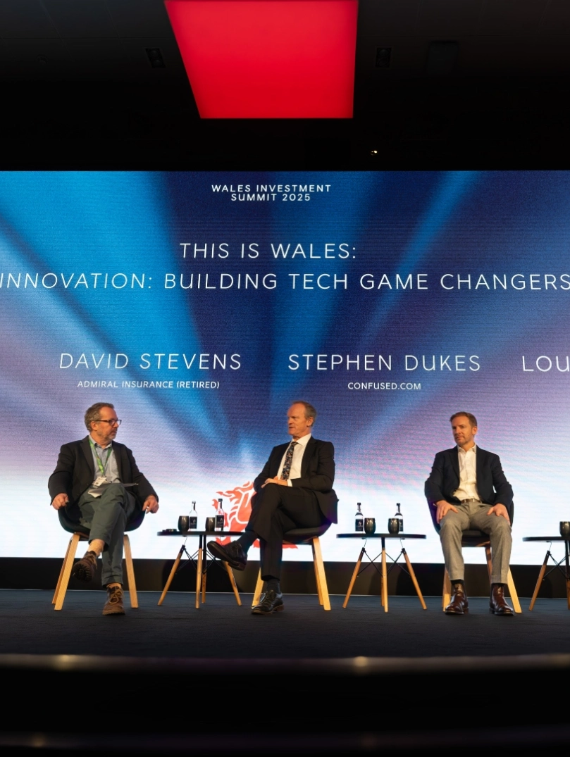 fPanel discussion on stage at a conference, with four speakers seated beneath a large screen reading “This is Wales: Investment to Innovation – Building Tech Game Changers from Wales.”