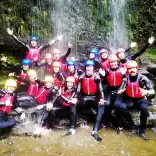 A group of people wearing wet suits, helmets and life jackets under a waterfall.