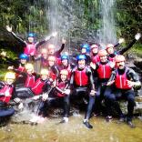 A group of people wearing wet suits, helmets and life jackets under a waterfall.