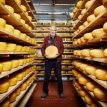 The owner holding a block of cheese surrounded by cheese on shelves.