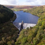 Aerial view of a river flowing over an open dam surrounded by trees and mountains.