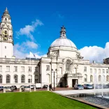 External of building white brick glass windows and clock on tower, with fountain and lawn green in the forefront.