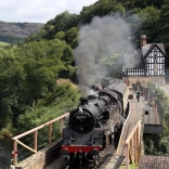 A steam train pulling out of a station alongside a lake.