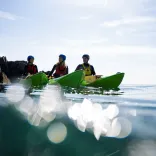Three kayakers on the ocean with a rocky cove backdrop.