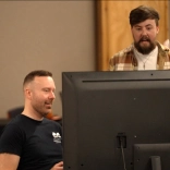Ambassador Richard Hebblewhte sitting at a computer screen talking with a collegue standing behind him in a checked shirt 