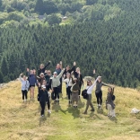 Group of people standing on a grassy hilltop with arms raised, overlooking a vast forest of evergreen trees and scattered countryside houses in the distance
