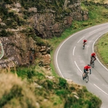 Cyclists riding along a winding mountain road in Wales with a painted rock face beside them.