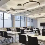 Meeting room featuring modern tables and chairs, large windows with city views, and circular ceiling lights, set up for a professional corporate event or training session.