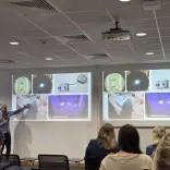 Presenter pointing to projected slides showing close‑up technical images, with an audience seated in a lecture room