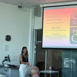 Presenter standing beside a screen showing a Cardiff University slide during a talk in a modern meeting room.