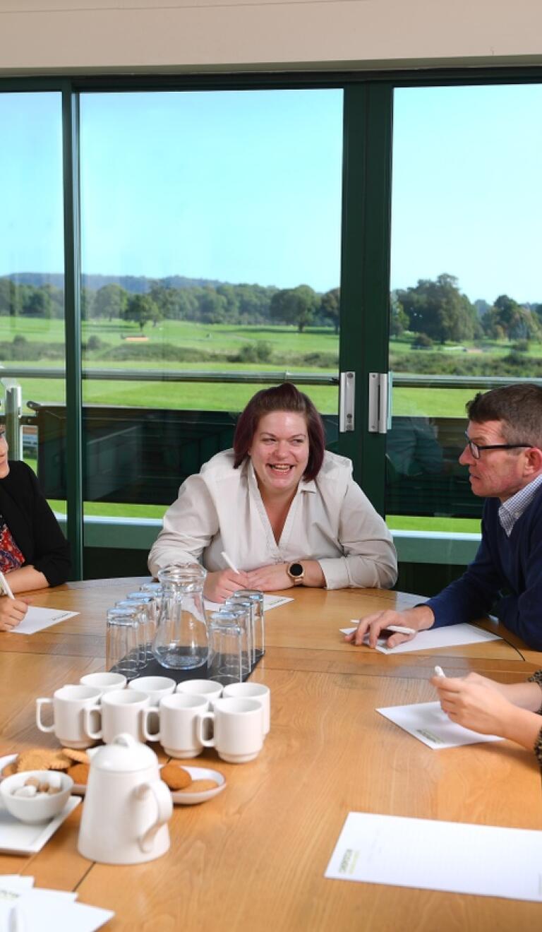 People around a meeting table with views of a racecourse beyond.