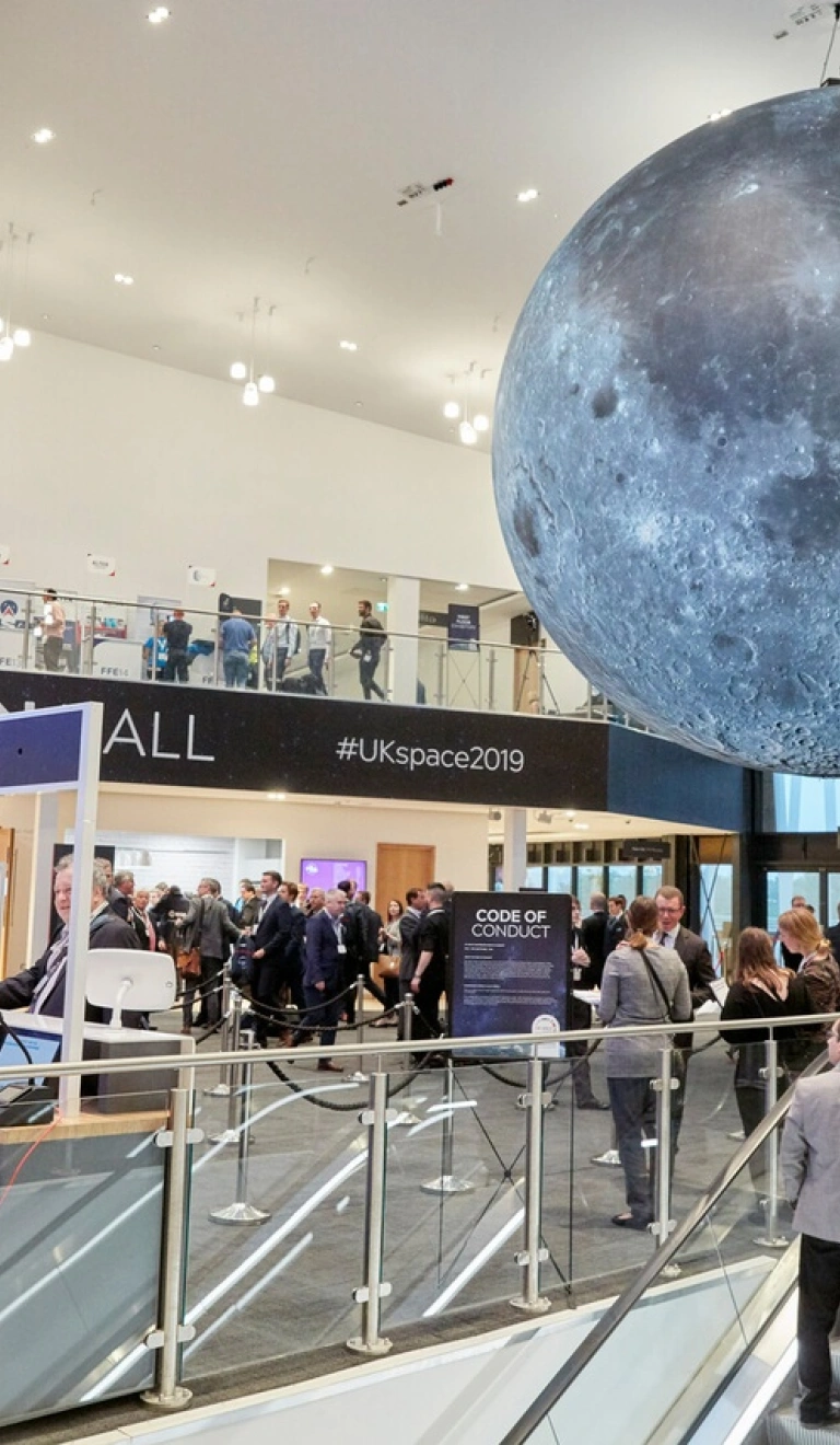 Exhibition hall with people coming up esculators to reception desks and a large grey space globe suspended from the ceiling.