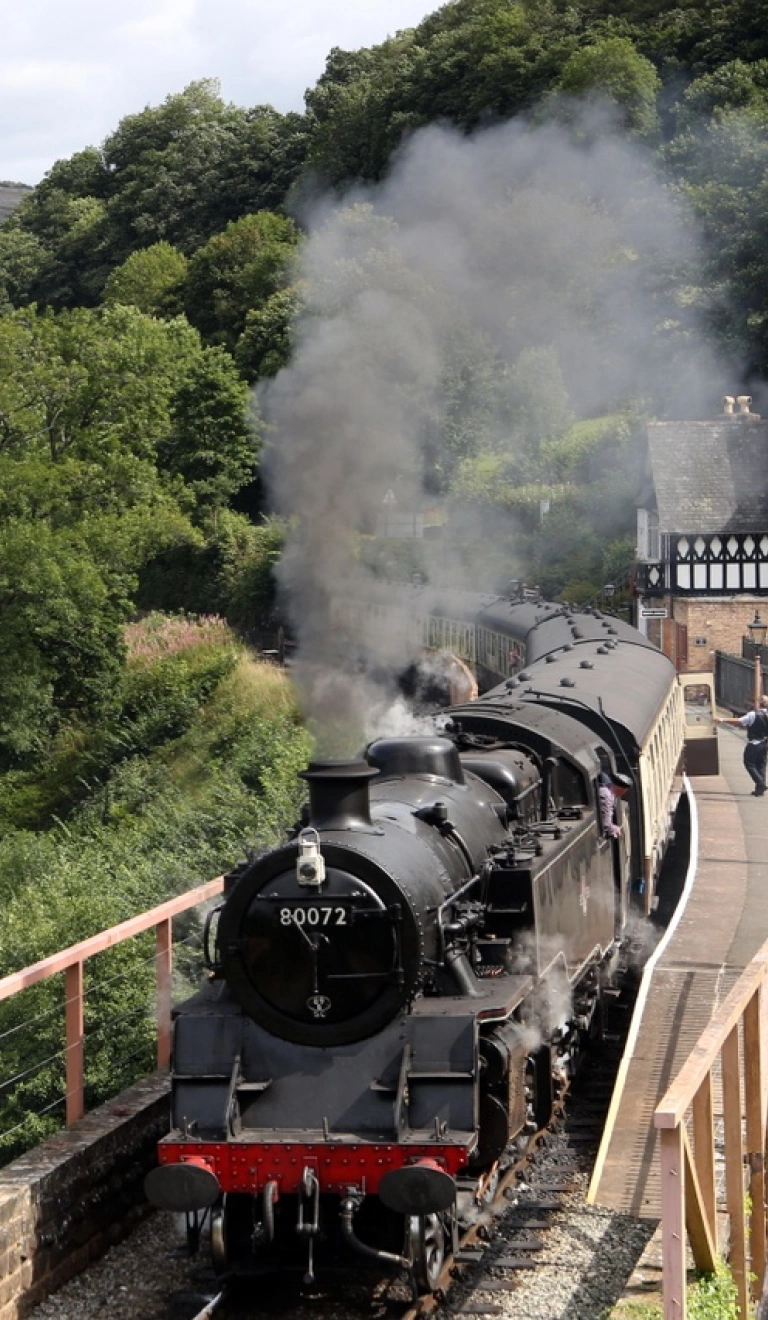 A steam train pulling out of a station alongside a lake.