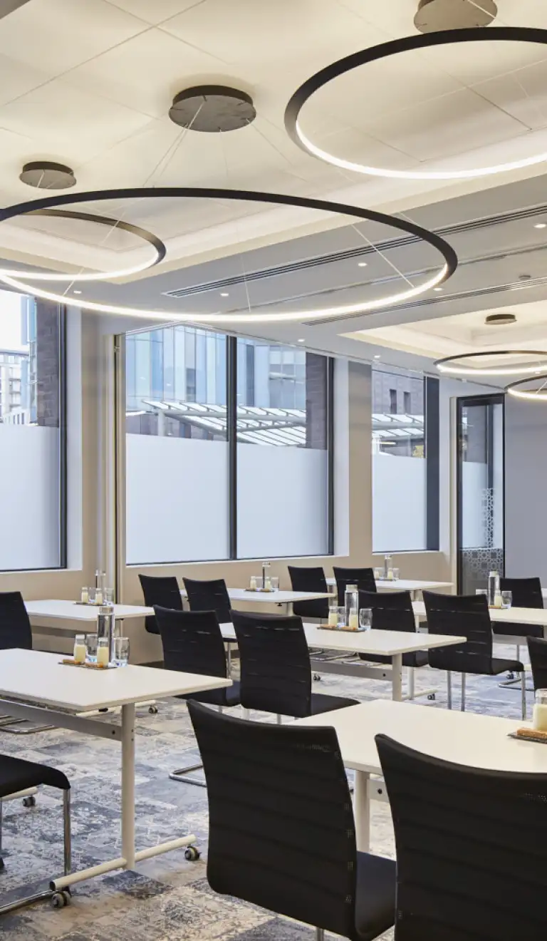 Meeting room featuring modern tables and chairs, large windows with city views, and circular ceiling lights, set up for a professional corporate event or training session.